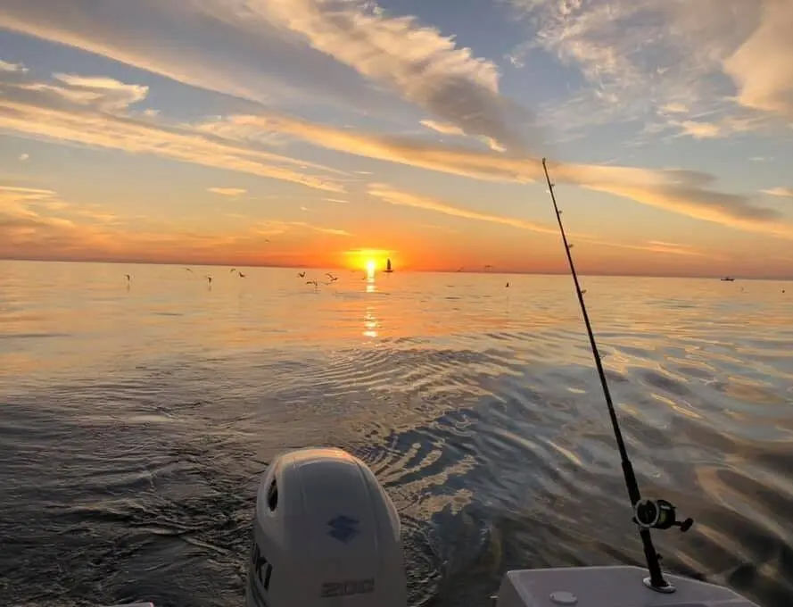 A fishing pole hangs of the stern of a boat in front of a sunset.